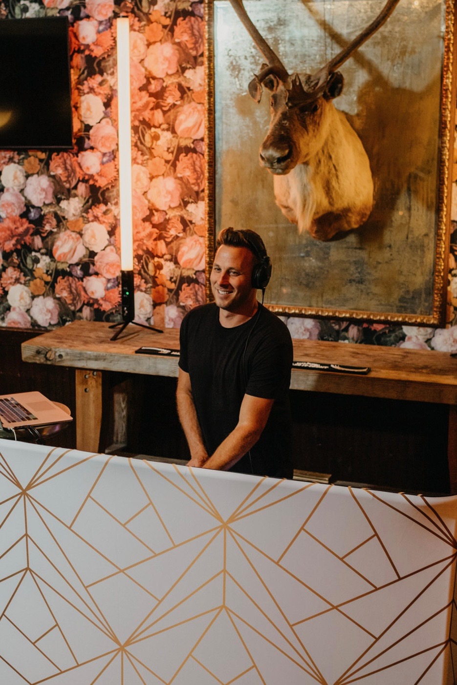 DJ Nick Dean wearing headphones behind his booth and its black and gold geometric facade