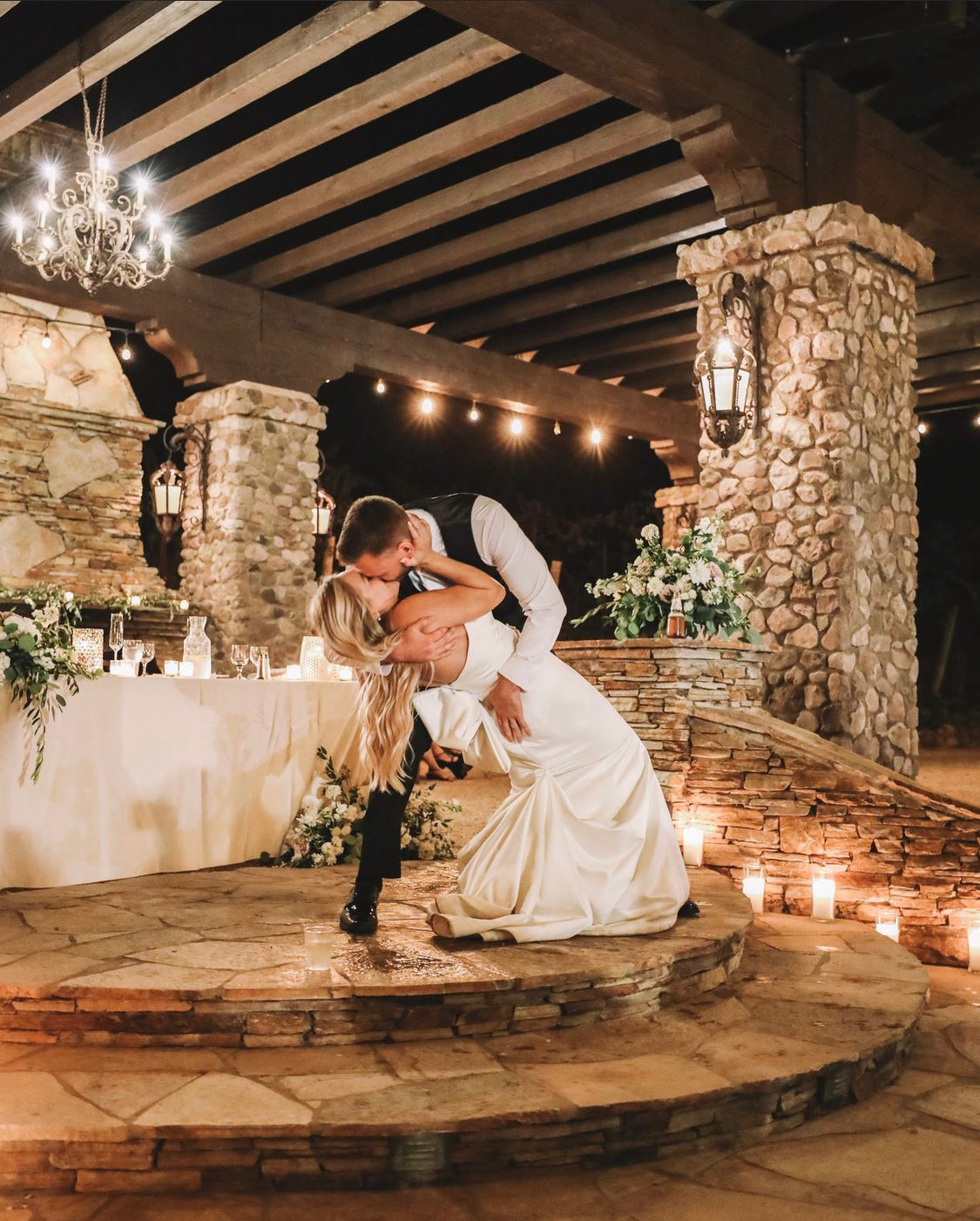 A newlywed couple share a dip kiss on a candlelit stone dance floor
